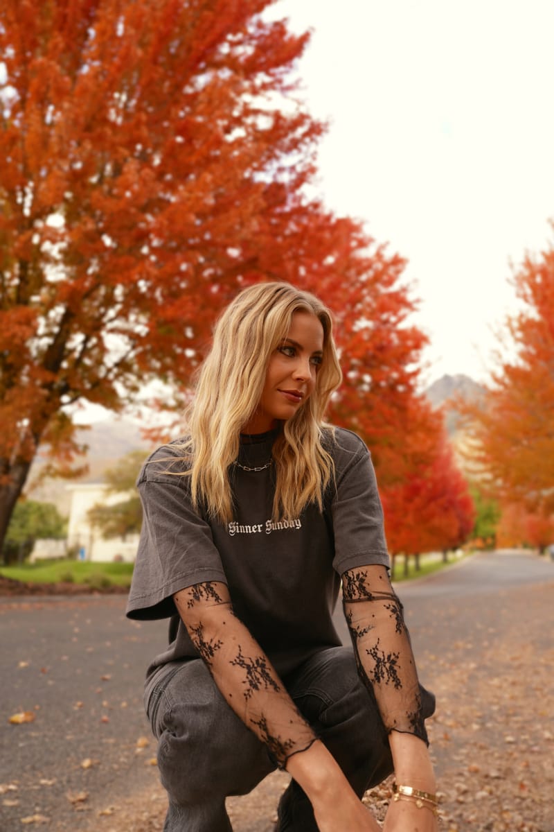 The Saints & Sinners tee worn crouching on a leaf-strewn autumn street over black lace sleeves, SINNER SUNDAY in small white blackletter across the chest
