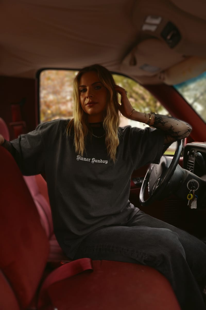 The Saints & Sinners tee worn in the cab of a red-interior pickup, SINNER SUNDAY printed small across the chest